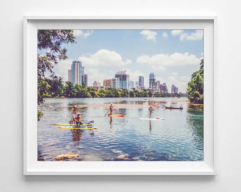 K&ouml;nnte beinhalten: Ein Panoramablick auf eine Skyline mit mehreren Geb&auml;uden, darunter ein hoher Wolkenkratzer, die sich im Wasser spiegeln. Menschen paddeln auf dem Wasser vor der Stadt.