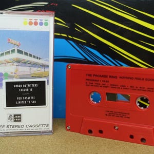 May include: A red cassette tape and a white cassette case. The case has a colorful design and text that reads "URBAN OUTFITTERS EXCLUSIVE RED CASSETTE LIMITED TO 500". The red cassette tape has the text "THE PROMISE RING NOTHING FEELS GOOD".