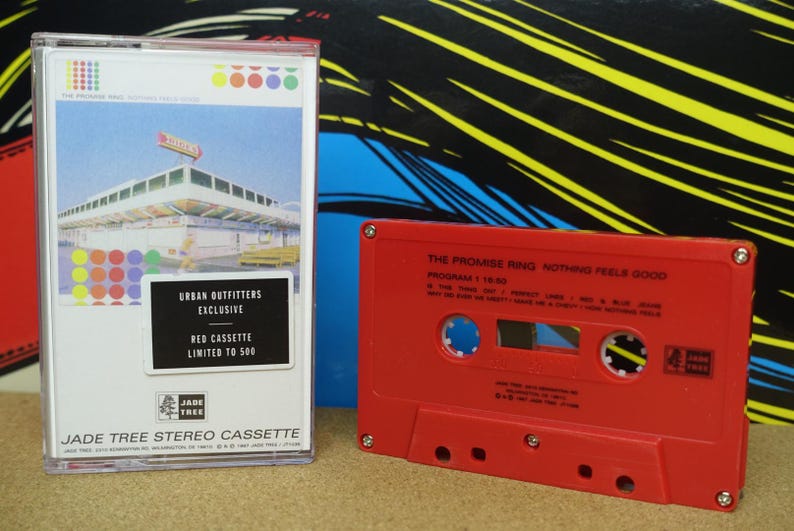 May include: A red cassette tape and a white cassette case. The case has a colorful design and text that reads "URBAN OUTFITTERS EXCLUSIVE RED CASSETTE LIMITED TO 500". The red cassette tape has the text "THE PROMISE RING NOTHING FEELS GOOD".