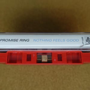 May include: A vintage cassette tape with a red case and a white label. The label reads "THE PROMISE RING" and "NOTHING FEELS GOOD" in blue text. The cassette is labeled "JADE TREE" and "JT1036".