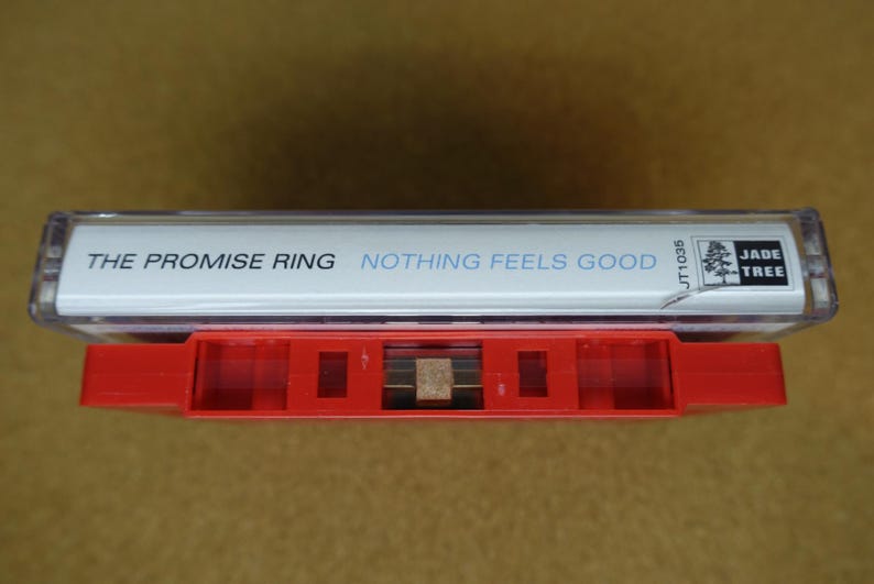 May include: A vintage cassette tape with a red case and a white label. The label reads "THE PROMISE RING" and "NOTHING FEELS GOOD" in blue text. The cassette is labeled "JADE TREE" and "JT1036".