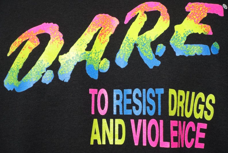 May include: Black fabric with the D.A.R.E. logo in neon colors. The text "TO RESIST DRUGS AND VIOLENCE" is below the logo, also in neon colors. The design has a retro, 80s or 90s aesthetic.