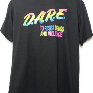 May include: Black t-shirt with the D.A.R.E. logo in rainbow colors. Below the logo, the text reads "TO RESIST DRUGS AND VIOLENCE" in a similar rainbow gradient. The shirt is displayed on a mannequin.