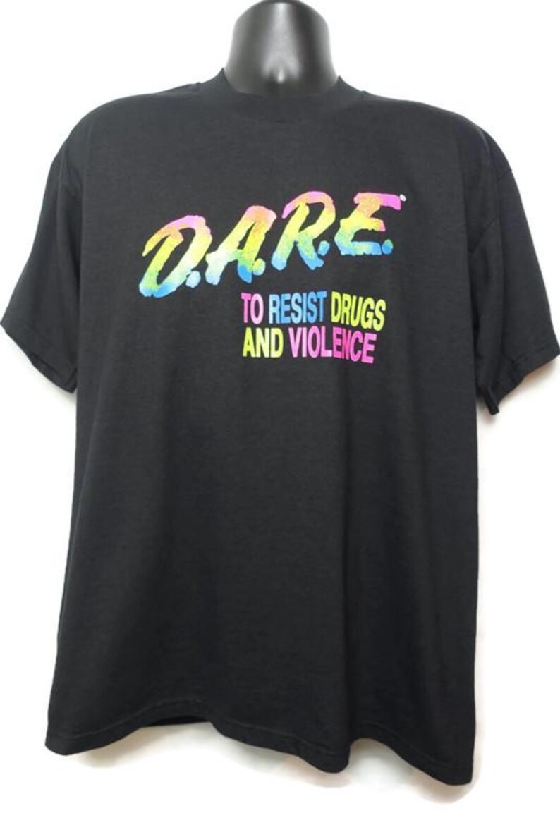 May include: Black t-shirt with the D.A.R.E. logo in rainbow colors. Below the logo, the text reads "TO RESIST DRUGS AND VIOLENCE" in a similar rainbow gradient. The shirt is displayed on a mannequin.