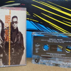 Könnte beinhalten: Eine Vintage-Kassette des Albums "Inner City - Big Fun". Das Kassettenetui zeigt ein Foto eines Mannes und einer Frau. Die Kassette ist transparent mit einem schwarzen Etikett und der Titelliste. Das Album wurde von Kevin Saunderson produziert.