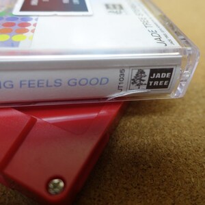 May include: A close-up of a cassette tape case with the text "NOTHING FEELS GOOD" in blue. The case is clear plastic, and the label features colorful circles and the "JADE TREE" logo. The tape is resting on a red plastic device.