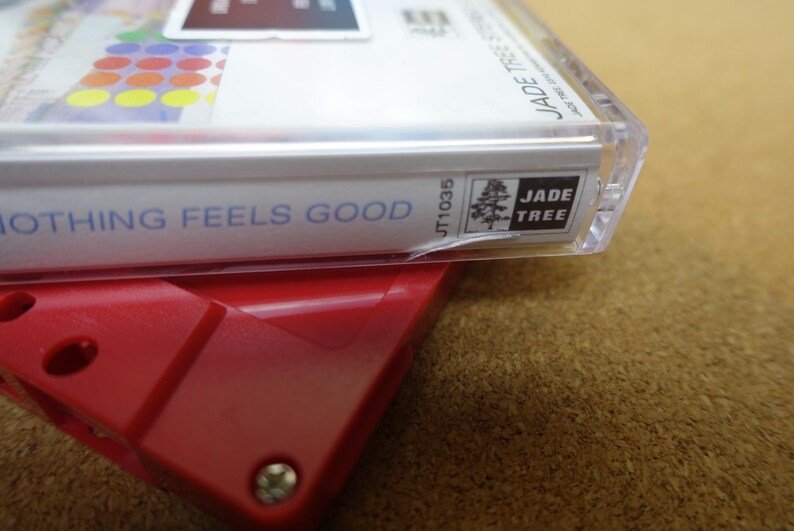 May include: A close-up of a cassette tape case with the text "NOTHING FEELS GOOD" in blue. The case is clear plastic, and the label features colorful circles and the "JADE TREE" logo. The tape is resting on a red plastic device.