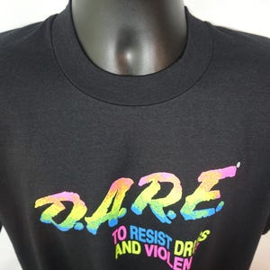 May include: Black crew neck t-shirt with the word "DARE" in a rainbow gradient font. Below the word "DARE" is the text "TO RESIST DRUGS AND VIOLENCE" in a white font. The shirt is displayed on a mannequin.