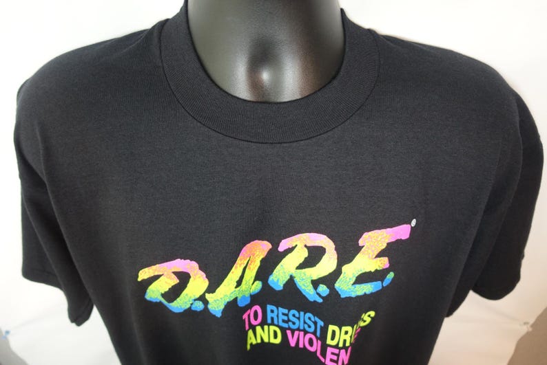 May include: Black crew neck t-shirt with the word "DARE" in a rainbow gradient font. Below the word "DARE" is the text "TO RESIST DRUGS AND VIOLENCE" in a white font. The shirt is displayed on a mannequin.