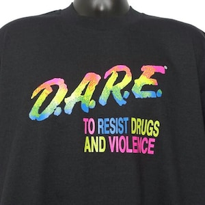May include: Black t-shirt with the word "DARE" in a colorful, rainbow-like font. Below the word "DARE" is the text "TO RESIST DRUGS AND VIOLENCE" in a mix of bright colors. The shirt is displayed on a black mannequin.
