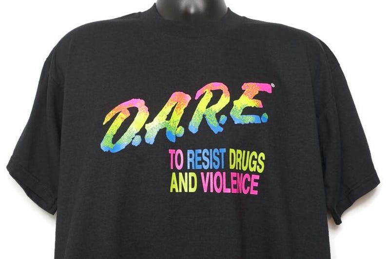 May include: Black t-shirt with the word "DARE" in a colorful, rainbow-like font. Below the word "DARE" is the text "TO RESIST DRUGS AND VIOLENCE" in a mix of bright colors. The shirt is displayed on a black mannequin.
