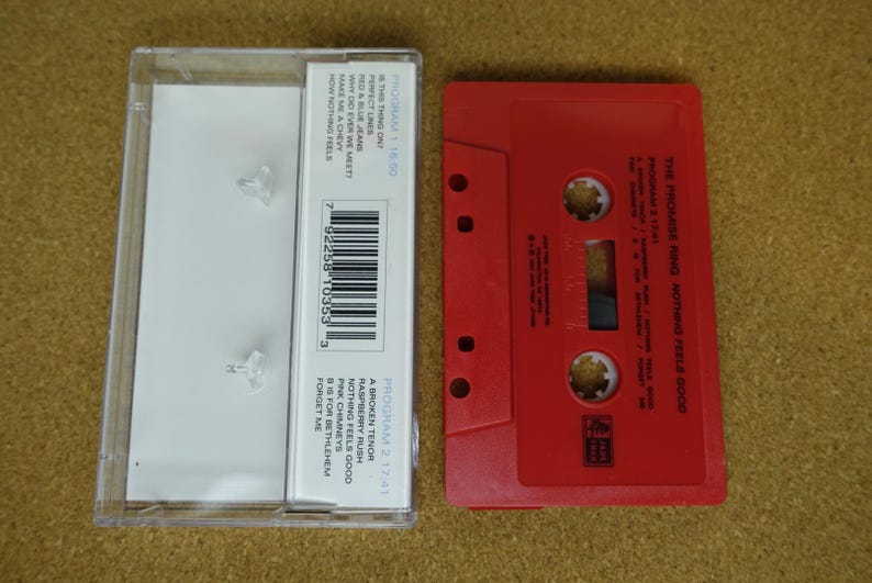 May include: A red cassette tape and its clear plastic case are displayed. The cassette tape is labeled "THE PROMISE RING NOTHING FEELS GOOD." The case has a tracklist and a barcode. The cassette is a vintage music format.