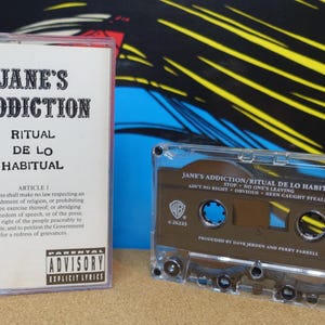 May include: A cassette tape and its case featuring the album "Ritual de lo Habitual" by Jane's Addiction. The case has the album title and lyrics. The cassette tape is transparent with a brown interior.