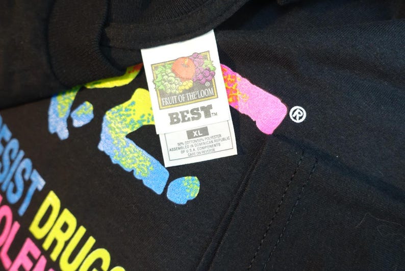 May include: Black t-shirt with colorful text that reads "RESIST DRUGS VIOLENCE". The shirt has a Fruit of the Loom tag that says "BEST" and "XL". The shirt has a colorful design.
