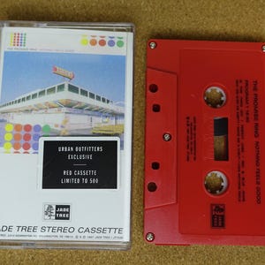 May include: A red cassette tape and its case are displayed. The case features a colorful design and the text "URBAN OUTFITTERS EXCLUSIVE RED CASSETTE LIMITED TO 500." The red cassette tape is labeled "THE PROMISE RING NOTHING FEELS GOOD."