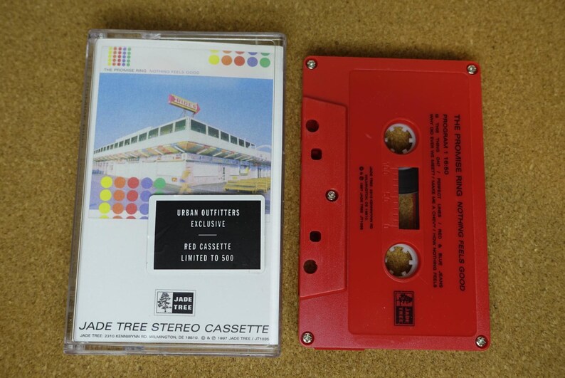 May include: A red cassette tape and its case are displayed. The case features a colorful design and the text "URBAN OUTFITTERS EXCLUSIVE RED CASSETTE LIMITED TO 500." The red cassette tape is labeled "THE PROMISE RING NOTHING FEELS GOOD."