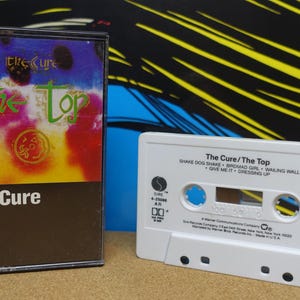 May include: A vintage cassette tape and case for "The Cure - The Top" album. The case has a colorful design with the band's name and album title. The white cassette tape has the album title and track list.