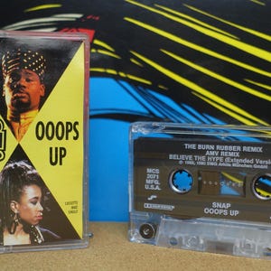 Könnte beinhalten: Zwei Musikkassetten. Die erste hat einen gelben Hintergrund mit dem Wort "SNAP!" und dem Text "OOOPS UP". Die zweite Kassette ist transparent mit schwarzem Text "THE BURN RUBBER REMIX" und "BELIEVE THE HYPE".