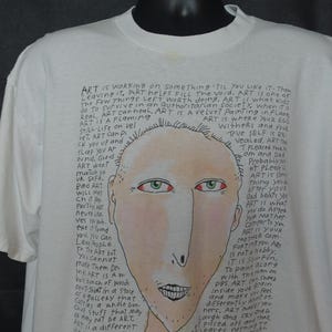 May include: White t-shirt with a hand-drawn illustration of a face and handwritten text. The face has red eyes and a long nose. The text includes phrases like "ART is where your ego withers" and "ART is a different thing."