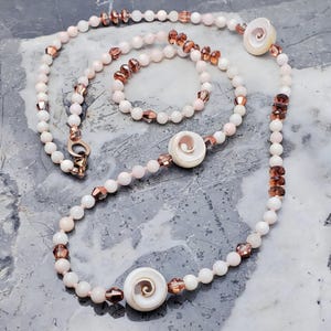 May include: A delicate necklace and bracelet set featuring pale pink beads and copper-colored accents. The necklace includes three white spiral-shaped pendants. The set is displayed on a gray surface.
