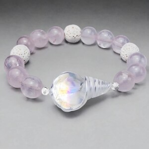 May include: A bracelet featuring light purple, round beads interspersed with white, textured beads. The bracelet has a clear, iridescent seashell-shaped clasp. The beads are approximately 0.5 inches in diameter.
