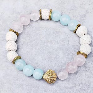Peut inclure: Un bracelet avec des perles de roche volcanique blanche, des perles bleu clair et rose, et un charme en forme de coquillage doré.