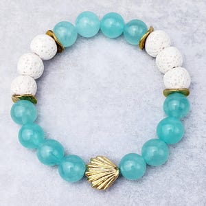 Pode incluir: Um bracelete de contas turquesa e brancas com um fecho de concha dourada. O bracelete é feito com contas de lava e jade.