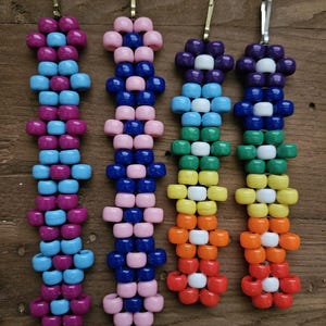 May include: Four vertical beaded keychains featuring flower patterns. The first is purple and light blue, the second is pink and dark blue, and the last two feature a rainbow color sequence with white centers, all attached to metal clips.
