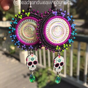 May include: A pair of beaded earrings with a colorful design and a sugar skull charm. The earrings are made with black, pink, blue, green, and white beads. The sugar skull charms are white with black and pink details. The earrings are hanging from silver hooks.