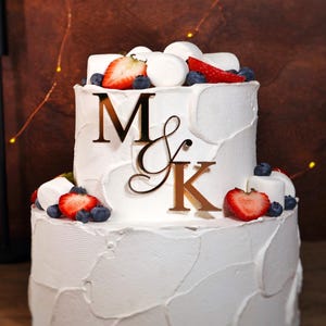 May include: A two-tiered white cake with textured frosting, decorated with fresh strawberries, blueberries, and marshmallows. A gold-colored monogram with the letters "M & K" sits on the front. The cake is approximately 4 inches by 4 inches.