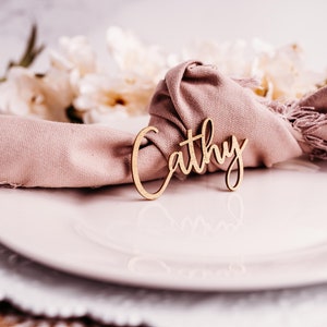 Può includere: Un segnaposto in legno con il nome "Cathy" scritto in corsivo. Il segnaposto è su un tovagliolo rosa, che è su un piatto bianco.