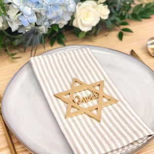 May include: A gold star of David place card with the name "David" cut out of the center. The place card is on a white and beige striped napkin on a white plate.