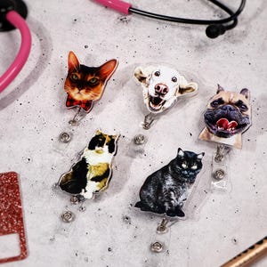 May include: A collection of pet-themed badge reels featuring cat and dog faces. The reels include a cat with a bow tie, a smiling dog, a bulldog, a calico cat, and a black cat. A pink stethoscope and a name tag are also visible.
