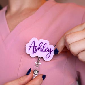 Personalized name badge reel worn on pink scrubs, showing elegant acrylic name tag design with 360-degree alligator clip and 25-inch retractable cord. Custom nurse badge reel with pastel acrylic base and glitter text, perfect for medical staff, CNAs, or teachers. Nurse week gift idea, handmade in the USA. Stylish retractable ID holder for nurses and healthcare professionals.