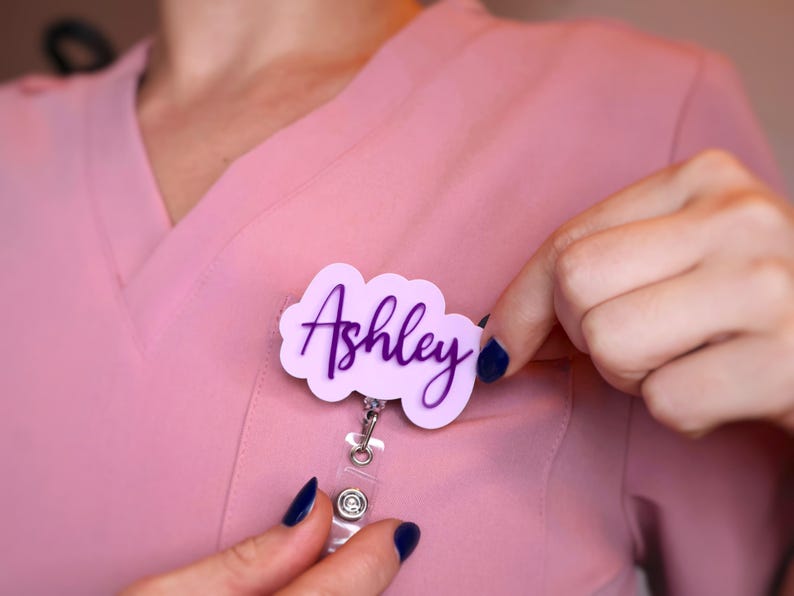 Personalized name badge reel worn on pink scrubs, showing elegant acrylic name tag design with 360-degree alligator clip and 25-inch retractable cord. Custom nurse badge reel with pastel acrylic base and glitter text, perfect for medical staff, CNAs, or teachers. Nurse week gift idea, handmade in the USA. Stylish retractable ID holder for nurses and healthcare professionals.