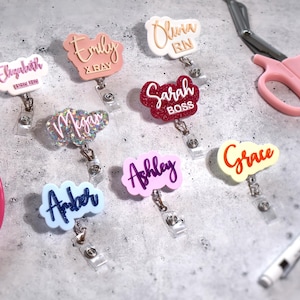 May include: A collection of name badge reels in various colors and designs. Each badge features a name in a unique font, such as "Elizabeth," "Emily," and "Olivia." Other items include pink medical scissors, a pen, and a stethoscope.