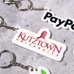 May include: Keychains with various designs, including the Kutztown University logo in red and white, and the PayPal logo in black and white. Each keychain has a silver chain and ring.
