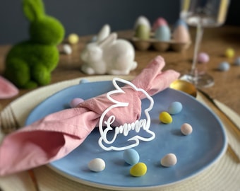 May include: A white bunny-shaped napkin ring with the name "Emma" written on it. The ring is holding a pink napkin and is placed on a blue plate with pastel-colored candy eggs.