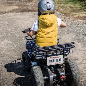 May include: A child wearing a yellow vest and a silver helmet is riding a small ATV on a dirt path. The ATV has a black rack and a license plate that reads "YOUR TEXT".