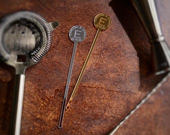 May include: Two silver cocktail stirrers with round, gold-colored tops. The tops have the text "CAROLINE & JOSEPH" and the letter "E" on them. The stirrers are on a brown, textured surface.