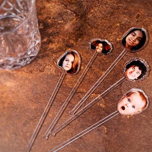 May include: A collection of clear plastic drink stirrers, each topped with a different person's face. The faces are of various ages and ethnicities. A decorative glass is in the background. The stirrers are on a brown surface.