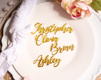 May include: Gold acrylic place cards with names Christopher, Olivia, Brian, and Ashley, arranged on a white plate with a white napkin and floral accents. Ideal for weddings or special events.