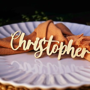 Può includere: Un segnaposto in legno con il nome "Christopher" scritto in corsivo. Il segnaposto è su un piatto bianco con un tovagliolo marrone.