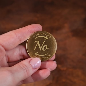 May include: A gold-colored decision coin with the word "No" in a stylized font. The coin also features the text "DECISION COIN" and "FLIP TO DECIDE" around the edges. The coin is held in a hand.