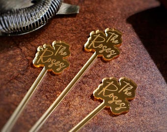 May include: Three gold-colored cocktail stirrers with a clover-shaped design. Each stirrer has the text "The Roses" written on it.