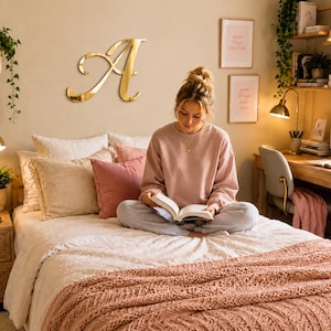 May include: A large, gold-colored, cursive letter A wall decoration mounted on a beige wall above a bed. The letter has a reflective, metallic finish and a flowing, elegant script font style, serving as a prominent focal point in the room.