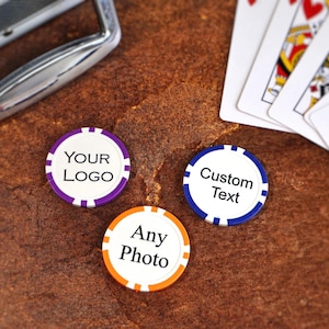 May include: Three poker chips with customizable text and logos, in purple, blue, and orange, are displayed on a textured brown surface. Playing cards are visible in the background, along with a silver metal object. The chips are designed for personalization.