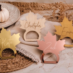 May include: Four maple leaf-shaped napkin rings in various finishes. One is light wood with the name Christopher, one is gold mirror with Kate, one is rose gold mirror with Zack, and one is brushed gold with the name Emma engraved in script.