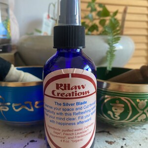 May include: A blue glass bottle with a black spray top and a white label that reads "RHaw Creations The Silver Blade Cleanse your space and Cut through negativity with this Refreshing scent. Feel your mind clear. Fill your space with happiness afterward. Ingredients: purified water, Dalmatian Sage*, French Lavender, Peppermint, and Frankincense 4 fl oz. "organic"
