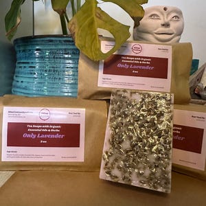May include: Two bars of handmade soap wrapped in brown paper. The soap is white with dried lavender flowers and the label reads "Tea Soaps with Organic Essential Oils & Herbs Only Lavender 3 oz".